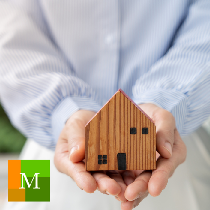 Person holding a wooden house model with a green and orange M logo